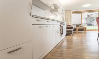 A modern kitchen with white cabinets and wooden floor. In the background, a cozy living area can be seen that looks bright and inviting.