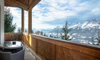 A beautiful balcony with a table and chairs offers a view of the mountains and the valleys. The sky is partly cloudy and there is a calm, peaceful atmosphere.