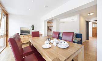 A modern dining room with a large wooden table and red chairs. In the background, bright walls and a television can be seen.