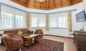 A cozy living room with a wooden ceiling and large windows. The furniture is comfortable and a television is mounted on the wall.