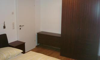 A simple bedroom with a bed, a nightstand, and a wooden wardrobe. The walls are light and the floor is made of wood.