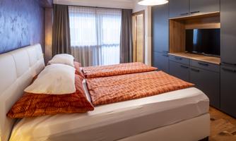 A modern bedroom with a double bed and orange bedspreads. Windows let natural light in and the room is well furnished.