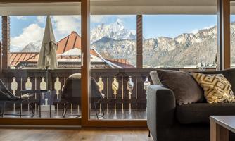 A cozy living space with a view of the mountains. Light flows in through large windows, and there is a balcony with chairs.