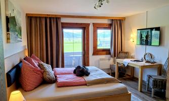 A cozy room with a large bed and checkered pillows. There is a table, chairs, and large windows that let in a lot of light.