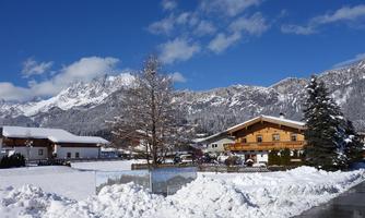 A picturesque winter village with snow-covered houses. In the background, majestic mountains rise under a clear blue sky.