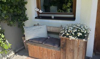 A cozy seating area made of wood with cushions and flowers. In the background, a large window reflects the surroundings.