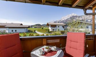 A cozy balcony with two red chairs and a table. In the background, green meadows and snow-covered mountains are visible.