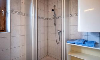 A modern shower with glass doors and tiled walls. On the left is a shelf with blue towels.