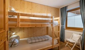 A cozy bedroom with a wooden bunk bed and a window. The walls are made of wood and there is a chair in the corner.
