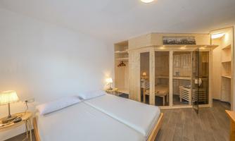 A modern bedroom with a double bed and a cozy atmosphere. On the right side, there is a wooden sauna and a small table with a lamp.