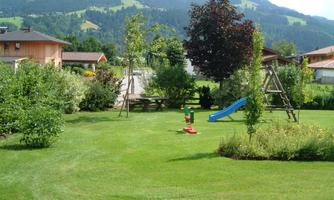 A beautiful garden with green grass, trees, and a playground. In the background, there are mountains and a quiet residential area.