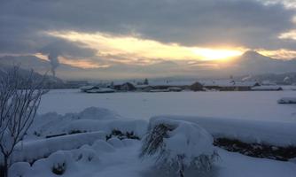 A snowy landscape with a gentle sunrise in the background. The sky is cloudy, and the surroundings are covered in fresh snow.
