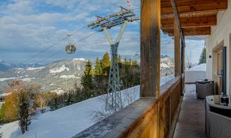 A view from a balcony of a snow-covered landscape. In the background, you can see a cable car running through the mountains.