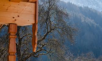 A view from a wooden balcony overlooking a picturesque mountain landscape. The sun shines over the mountains, and there are scattered trees visible in the foreground.