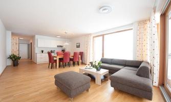 A modern living room with a sofa and a coffee table. In the background, there is a dining area with red chairs and a bright kitchen.
