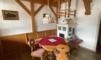 A cozy dining area with a round table and wood paneling. The room features a tiled stove and a rustic ceiling lamp.