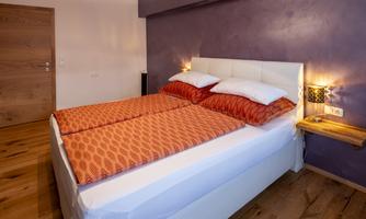A cozy bedroom with a large bed and orange bed covers. The walls are painted in a shade of purple and the floor is made of wood.