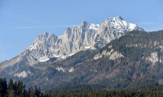 An impressive mountain panorama with snow-capped peaks and green forests in the foreground. The sky is clear and bright blue.