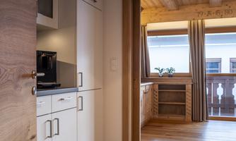 A modern kitchen with white cabinets and wooden details. In the background, a bright room with windows and plants can be seen.