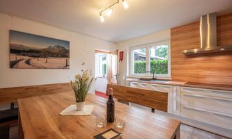 A modern kitchen with wood surfaces and plenty of natural light. On the table, there is a vase with flowers and a bottle.