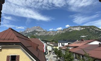 View of the mountains with green forests and blue sky. In the foreground, you can see roof tiles and small buildings.