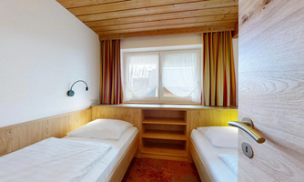 A cozy bedroom with two single beds and wooden paneling. Soft light filters into the room through the window.