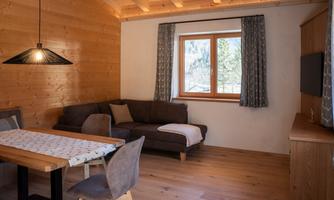 A cozy living space with wooden paneling and a large window. The room is equipped with a couch, a table, and chairs.