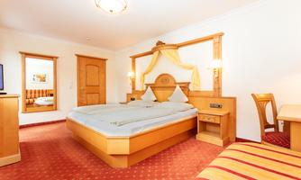 A cozy hotel room with a large double bed and wooden furniture. The walls are bright and the floor is covered with red carpet.