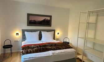 A bright bedroom with a large bed and white pillows. A picture hangs on the wall, and there is a modern shelf.