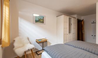 A cozy bedroom with two beds and a small table. The walls are light, and there is a window with yellow curtains that provide natural light.
