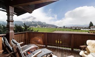 A cozy balcony with a wooden chair and a view of a green landscape and snow-covered mountains. The sky is partly cloudy and there is plenty of space for relaxation.