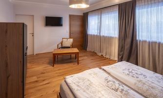 A cozy bedroom with two beds, a table, and a chair. Large windows provide plenty of daylight.