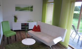 A modern furnished living room with a white sofa, a round table, and a green carpet. Large windows let in plenty of daylight and offer a view of the garden.