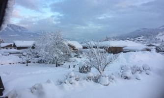 A snowy landscape with white hills and snow-covered trees. In the background, huts and mountains can be seen.