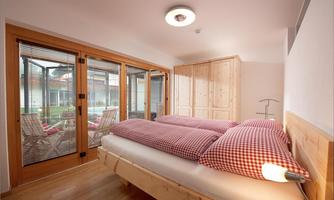 A cozy bedroom with a double bed and red-and-white checked pillows. Large windows offer a view of the garden and bring in plenty of daylight.