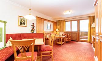 A cozy living area with a dining table and red upholstered furniture. Large windows provide access to a balcony and plenty of daylight.