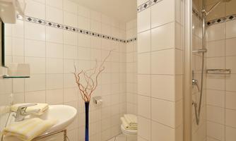 A modern bathroom with white tiles and a shower. A towel lies on the sink, and a decorative vase stands in the room.