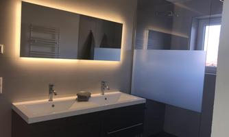 A modern bathroom with a double sink and a large mirror. The shower is separated by a frosted glass panel and there are illuminated walls.