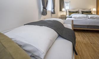 A modern bedroom with two beds. The walls are bright and the floors are made of wood, with curtains and lights in the background.