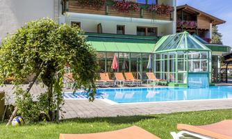 A beautiful pool area with loungers and a glass extension. In the background, there is a building with flower balconies.