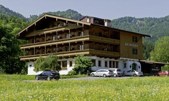 A traditional wooden building with several floors, surrounded by meadows and trees. In the foreground, some cars are parked.