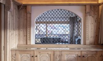 A cozy room with wooden paneled walls and a large window featuring a decorative lattice edge. A modern interior can be seen through the window.