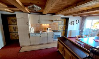 A modern kitchen with wooden shelves and a dining area.  
A lot of light shines in through the windows, and the floor is covered with a red carpet.