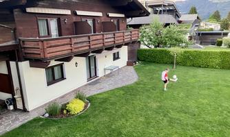 A house with wooden cladding and a large garden. A boy is playing with a ball on the lawn.