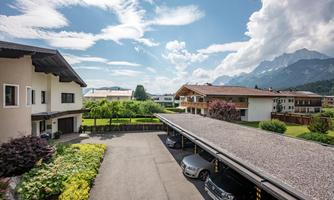 A quiet residential area with modern houses and green spaces. Majestic mountains rise in the background under a partly cloudy sky.