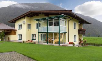 A modern two-story house with a winter garden. The green meadow and the mountains in the background create a serene atmosphere.