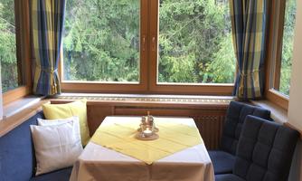A cozy dining area with a round table and seating. The large windows offer a view of green trees.