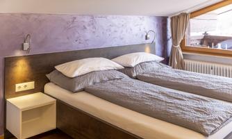 A modern bedroom with two single beds and a purple wall. Natural light streams through the window glass.