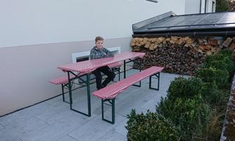 A boy is sitting at a long table with red and white tablecloths outdoors. In the background, wooden logs and trees are visible.