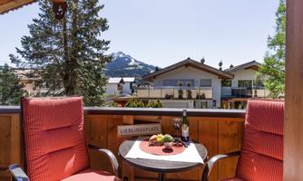 A cozy balcony with two red chairs and a small table. In the background, mountains and modern houses are visible.